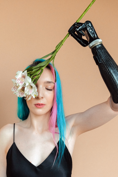 A model with a prosthetic arm and blue hair has one eye closed and is covering the other with a white flower. 