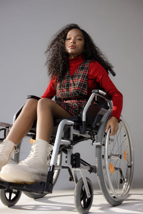 A Black girl in a wheelchair looks defiantly down at the camera in this picture. There is a plain grey background behind her.