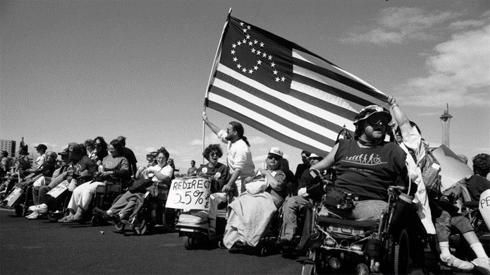 A long line of people in wheelchairs stand on top of a hill holding signs. One of them is waving an American flag but the stars are arranged to form a wheelchair. The picture is in black-and-white.